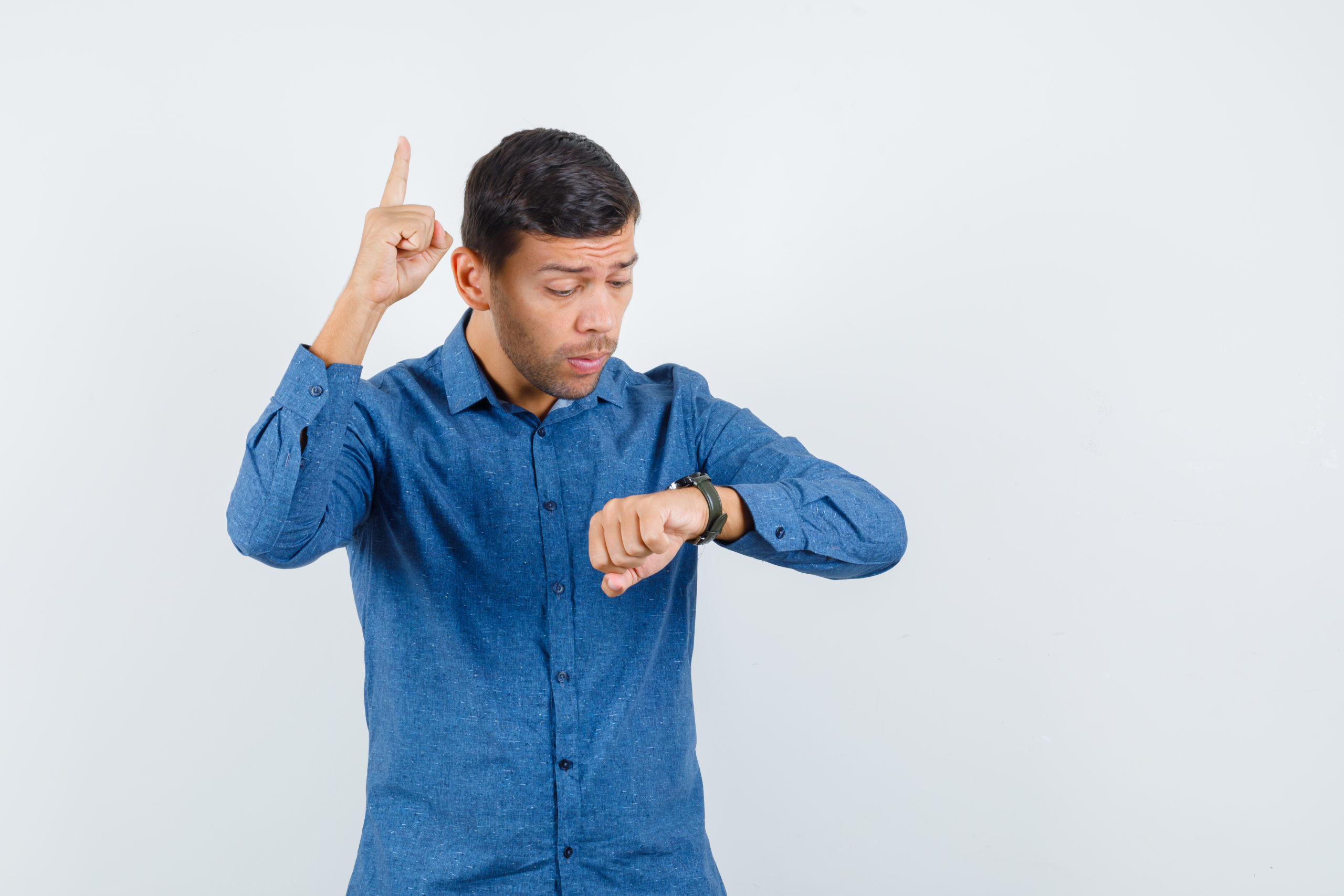Young Man Looking At His Watch With Finger Up In Blue Shirt And Looking Worried. Front View. - Contabilidade em Santa Catarina | Amaral Contabilidade