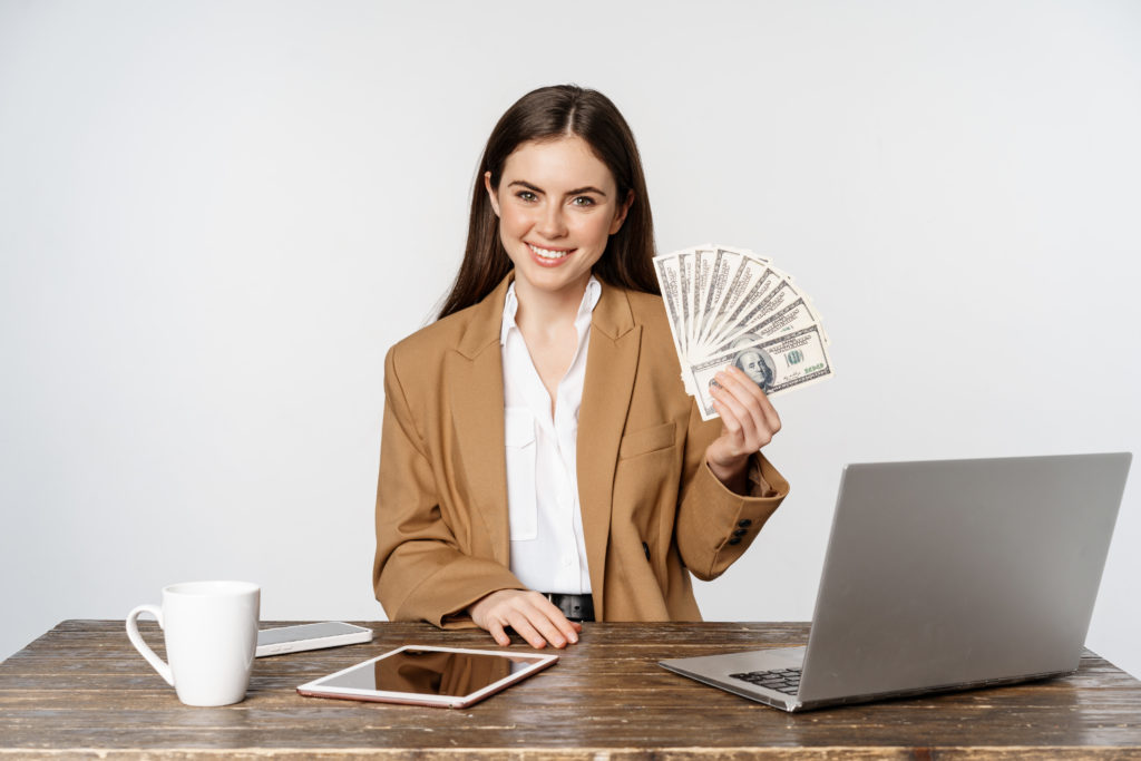 Portrait Of Businesswoman Sitting In Office With Money, Working And Making Profit Income, Posing Happy Against White Studio Background - Contabilidade em Santa Catarina | Amaral Contabilidade